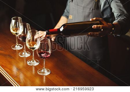 Bartender in apron pours sweet red wine from bottle in capacious glasses on wooden bar counter. Delicious alcohol drink in transparent glassware with nice scent.