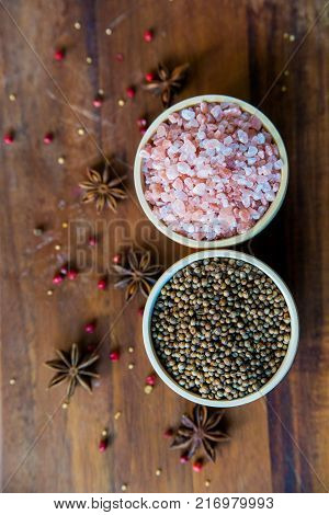 Pink Himalayan salt and coriander seeds in white bowls on wooden surface covered with anise and red peppercorn