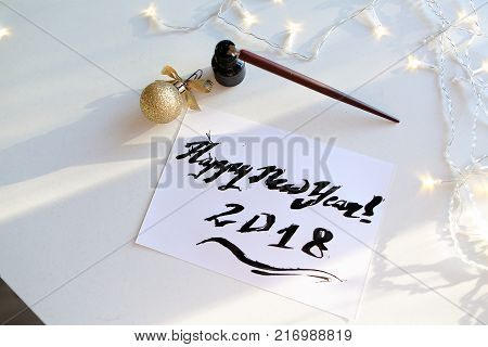 Christmas card with festive inscription Happy New Year, painted with black calligraphic font using carcass and fountain pen, which lies on table with Christmas garland in art workshop. Concept of beautiful writing, calligraphy and lettering, materials and
