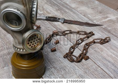 A gas mask stands next to a chain of rusty, scattered bullets and a knife on a wooden background