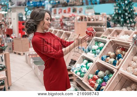 Cheerful happy customer girl choosing gifts for Christmas and New Year. She is taking red Christmas tree decorations. difficult decision. Various choice