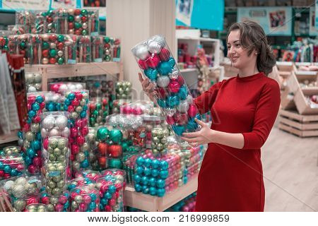 Cheerful happy customer girl choosing gifts for Christmas and New Year. She is taking red Christmas tree decorations. difficult decision. Various choice