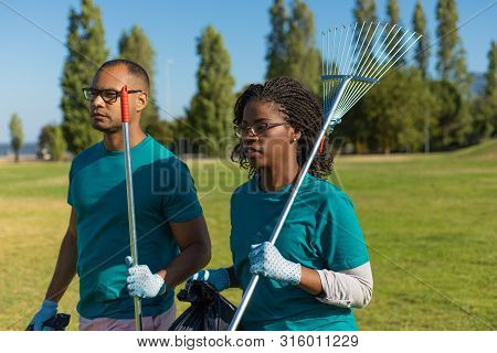 Couple Volunteers Working In Park. Black Woman And Latin Man Walking On Grass And Carrying Rakes And