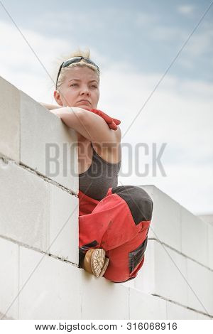Woman Wearing Dungarees Thinking About Future Of Building Her Home. Female Sitting On Wall Made Of A