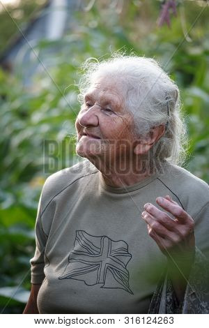 Close-up Portrait Of An Old Woman With Gray Hair Hangs Clothes To Dry On A Blurred Background Of A G