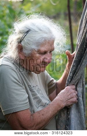 Close-up Portrait Of An Old Woman With Gray Hair Hangs Clothes To Dry On A Blurred Background Of A G