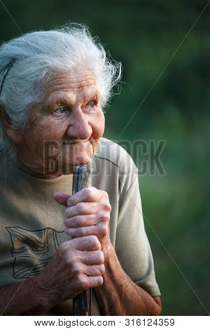 A Close-up Portrait Of An Old Woman With Gray Hair Smiling And Looking Up, Resting Her Chin On A Sti
