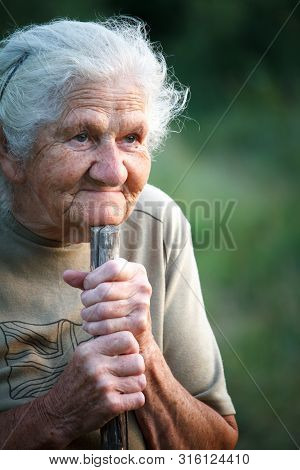 A Close-up Portrait Of An Old Woman With Gray Hair Smiling And Looking Up, Resting Her Chin On A Sti