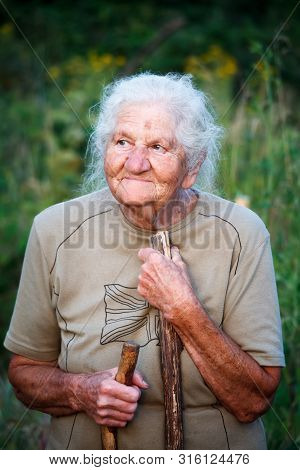 A Close-up Portrait Of An Old Woman With Gray Hair Smiling And Looking Up, Resting Her Chin On A Sti