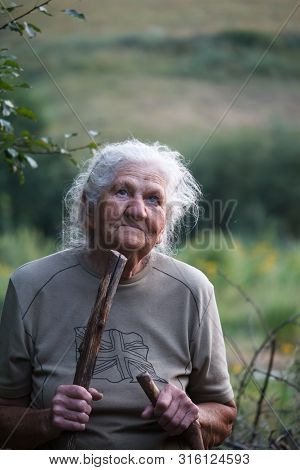 Portrait Of An Old Woman With Arthritic Feet Walking In A Grassy Meadow Leaning On A Stick As A Cane