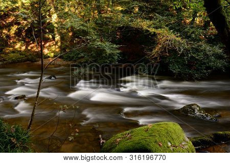 Long Exposure Of The River Flowing Through The Woods At Tarr Steps In Devon