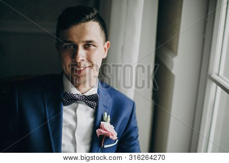 Stylish Happy Groom In Blue Suit, With Bow Tie And Boutonniere Posing And Smiling Near Window In Hot