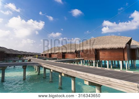 Luxury Hotel With A Line Of Bungalows Over The Water And A Wooden Path Between Them.