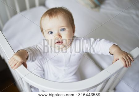 Adorable Baby Girl In Co-sleeper Crib Attached To Parents Bed. Little Kid Sitting In Cot