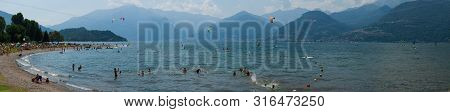 Lake Como, Italy - July 21, 2019: People Resting On Beach Near Lake. Trees And Alp Mountains On Back