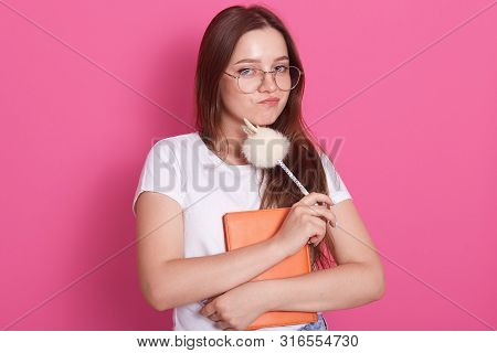 Horizontal Shot Of Young Woman With Staright Hair Holding Notebook And Pen While Standing Against Pi
