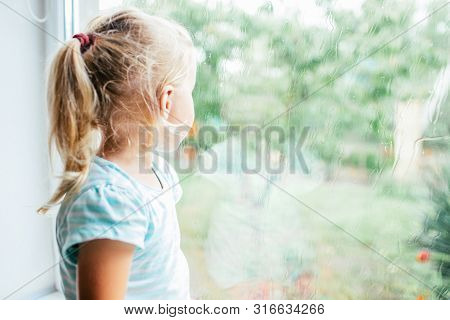 A Gorgeous Blonde Little Girl With Ponytale Staring Out Of The Window On A Wet, Cold Rainy Summer Da