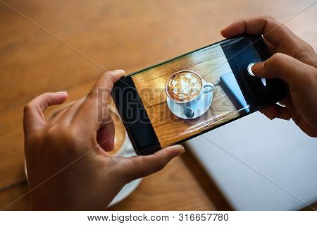 Hand Taking A Picture Of Hot Latte Art Coffee Cup With Art Foam On Wooden Table With Her Smart Phone