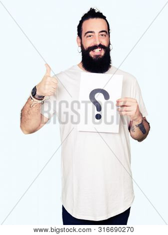 Young man with long hair and bear holding a paper with question mark happy with big smile doing ok sign, thumb up with fingers, excellent sign