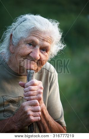 A Close-up Portrait Of An Old Woman With Gray Hair Smiling And Looking Up, Resting Her Chin On A Sti
