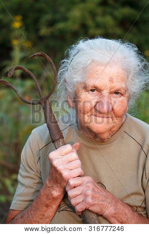 Close-up Portrait Of An Old Woman With Gray Hair Holding A Rusty Pitchfork Or Chopper In Her Hands,