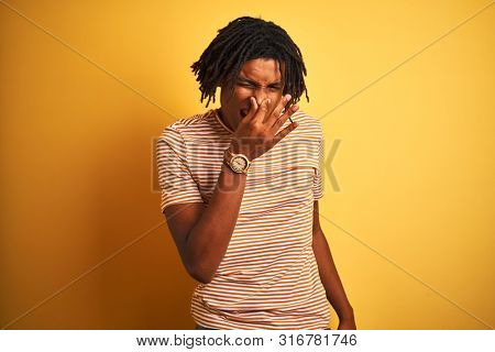 Afro man with dreadlocks wearing striped t-shirt standing over isolated yellow background smelling something stinky and disgusting, intolerable smell, holding breath with fingers on nose. Bad smells