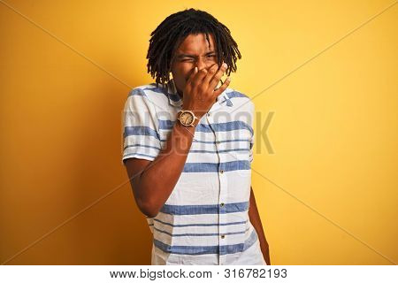 Afro american man with dreadlocks wearing striped shirt over isolated yellow background smelling something stinky and disgusting, intolerable smell, holding breath with fingers on nose. Bad smells