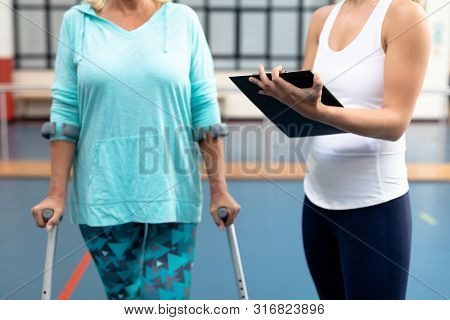 Mid section of female physiotherapist discussing with disabled senior woman on clipboard in sports center. Sports Rehab Centre with physiotherapists and patients working together towards healing