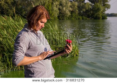 Female Ecologist Entering Information On Green Algae Taken In River On Tablet