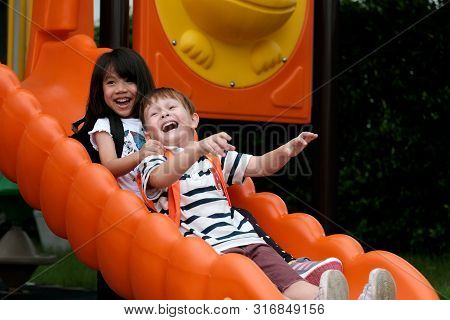 Happy Asian Girl And Caucasian Boy Playing Slide At School, Pre School Kids At Playground With Happi