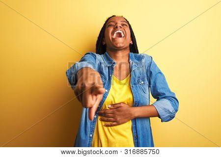 Young african american woman wearing denim shirt standing over isolated yellow background laughing at you, pointing finger to the camera with hand over body, shame expression