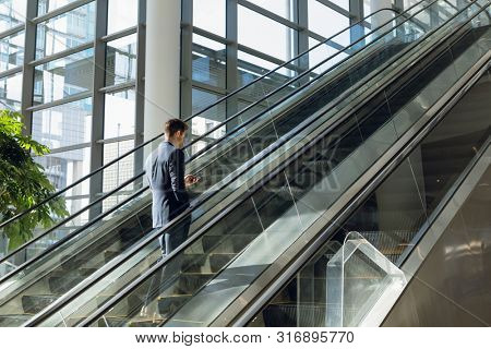 Full length side view of young Caucasian businessman going up an escalator in a modern office building using smartphone. Modern corporate start up new business concept with entrepreneur working hard