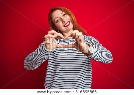 Young redhead woman wearing strapes navy shirt standing over red isolated background smiling in love doing heart symbol shape with hands. Romantic concept.