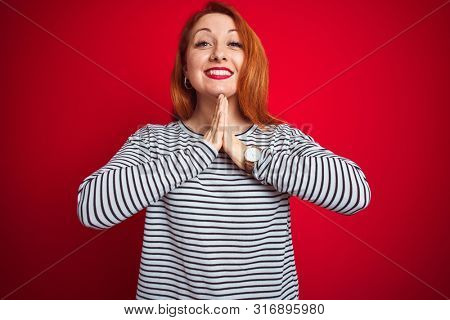Young redhead woman wearing strapes navy shirt standing over red isolated background praying with hands together asking for forgiveness smiling confident.