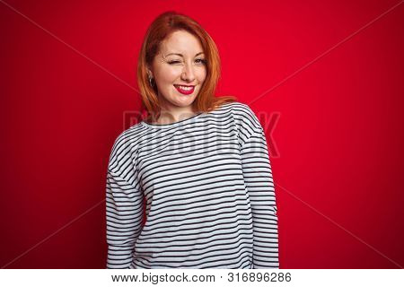 Young redhead woman wearing strapes navy shirt standing over red isolated background winking looking at the camera with sexy expression, cheerful and happy face.