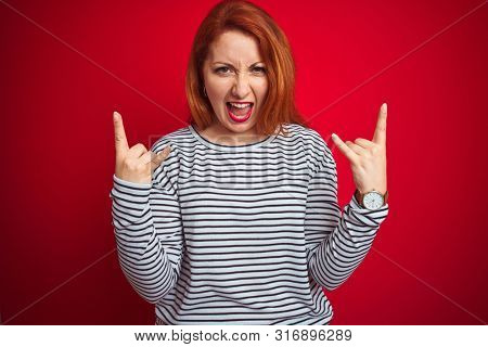 Young redhead woman wearing strapes navy shirt standing over red isolated background shouting with crazy expression doing rock symbol with hands up. Music star. Heavy concept.