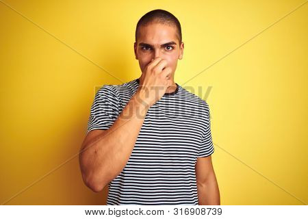 Young handsome man wearing striped t-shirt over yellow isolated background smelling something stinky and disgusting, intolerable smell, holding breath with fingers on nose. Bad smells concept.
