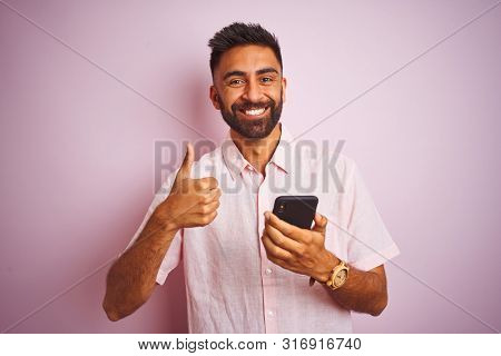 Young indian man using smartphone standing over isolated pink background happy with big smile doing ok sign, thumb up with fingers, excellent sign