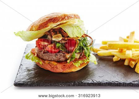 Fresh Beef Burger with Bacon, Fried Onions, Tomato Sauce, Pickled Cucumbers, Green Lettuce, Cheese and French Fries Garnish. Hamburger or Beefburger on Natural Black Stone Plate Isolated on White
