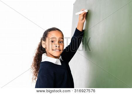 Cheerful African Amercan Schoolgirl Writing On Green Chalkboard While Smiling At Camera Isolated On 