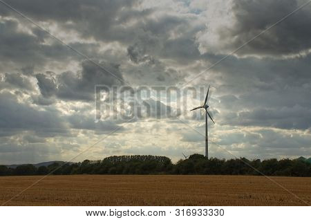 Wind Turbine In A Cloudy Sky Behind A Field And A Forest