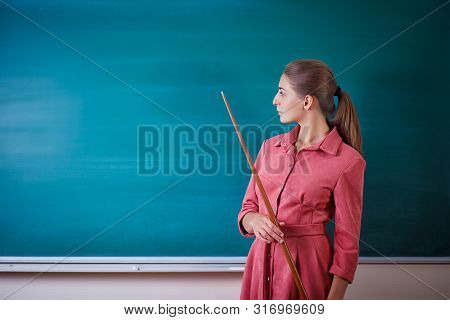 Young Woman Teacher Stands At A Blackboard With A Pointer. Template For The School Timetable And Oth