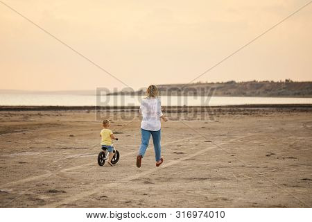 Mother With Son Riding Bicycle In Summer. Woman Runs Along With Kid On The Beach At Sunset. Son Lear