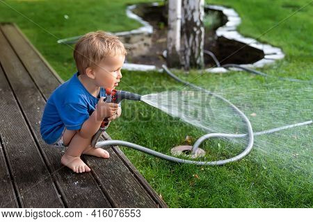 Cute Adorable Caucasian Blond Toddler Boy Enjoy Having Fun Watering Garden Flower And Lawn With Hose