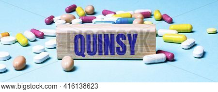 A Wooden Block With The Word Quinsy Stands On A Blue Background Among Multi-colored Pills. Medical C
