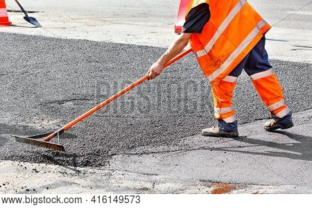 A Road Worker In A Bright Orange Overalls Renews A Section Of The Road With Fresh Hot Asphalt And Le
