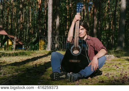 Portrait Of A Man With A Guitar In The Forest. Against The Background Of The Forest Camp.