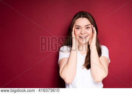 A Beautiful Girl In A White T-shirt With Long Hair Puts Her Hands To Her Face And Smiles Broadly In 