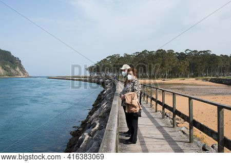 Aged Couple 70-80 Walking In Rodiles, Near The River And Puntal Forest In The Background, Asturias.