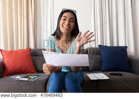 Mixed race gender fluid man working at home making video call holding documents, smiling and waving. staying at home in isolation during quarantine lockdown.